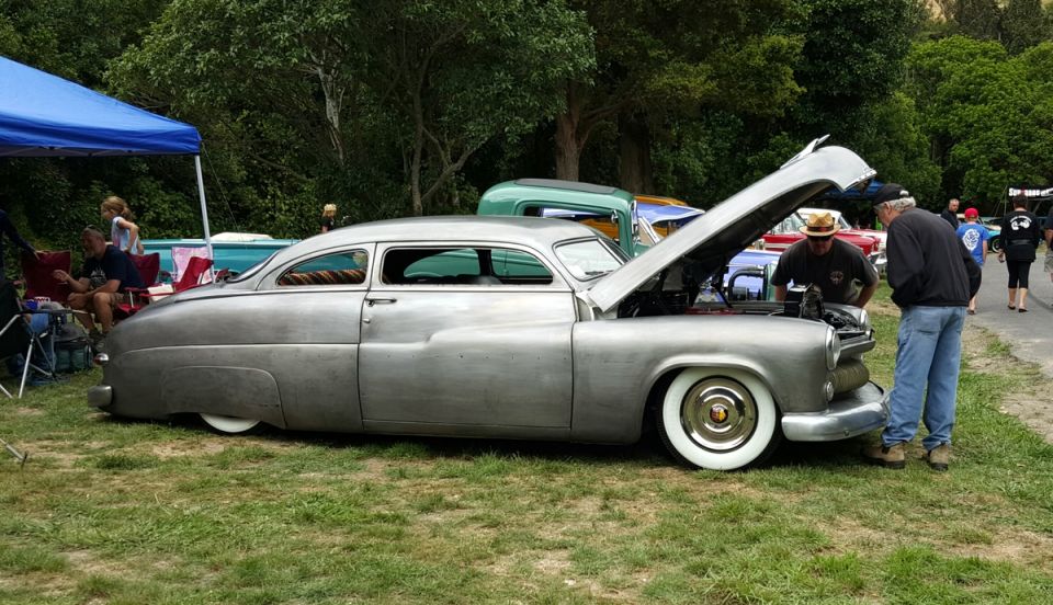 Custom silver 1950s sedan with hood open, two men looking at engine, other cars and people in background.