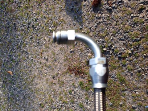 1979 Ford Bronco - Metal hose fitting with braided hose section against a gravel background.