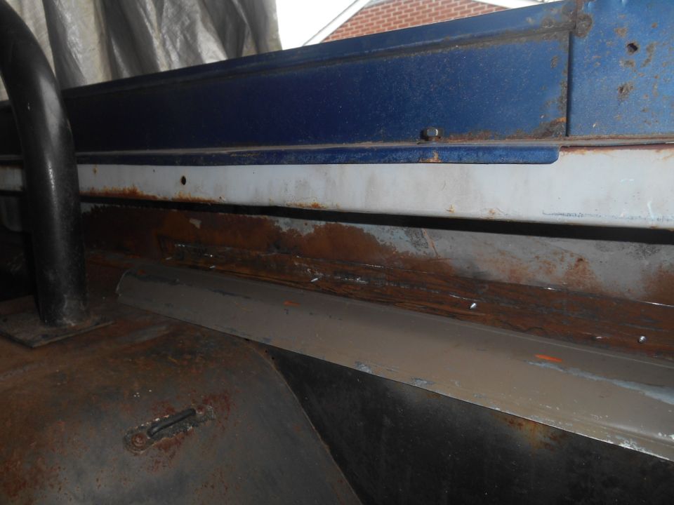 Close-up of the interior of a vehicle showing significant rust on the floor and lower body panels. A black roll bar is visible on the left