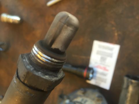1995 Ford Bronco - A close-up, angled shot shows the threaded end of a used oxygen sensor, with some carbon buildup visible on the ceramic tip. A white label…