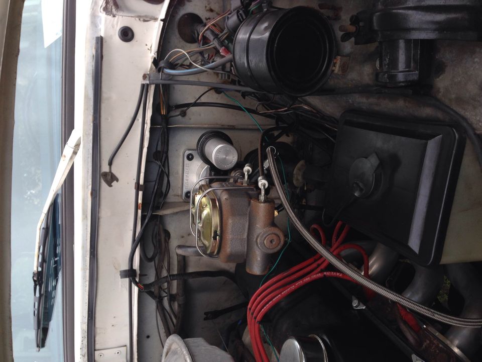 Engine bay of a classic Ford Bronco, showing brake master cylinder, red spark plug wires, and hoses.