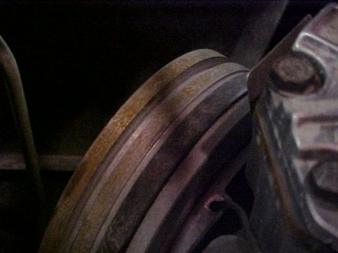 1995 Ford Bronco - Dim close-up of a vented brake rotor edge showing surface rust on the outer face and machined bands, with the caliper body at right. Shot f…
