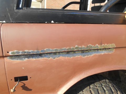 1972 Ford Bronco - A close-up of the rusted and repaired fender of a classic Ford Bronco. The image shows areas of exposed metal, primer, and what appears to…