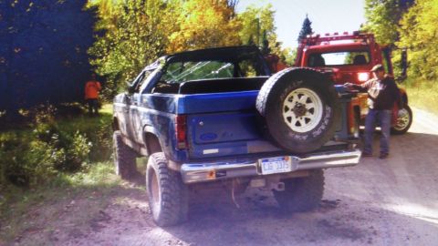 1985 Ford Bronco - A blue Ford Bronco with a broken rear window is parked on a dirt road next to a red tow truck. A person in an orange shirt stands in the ba…