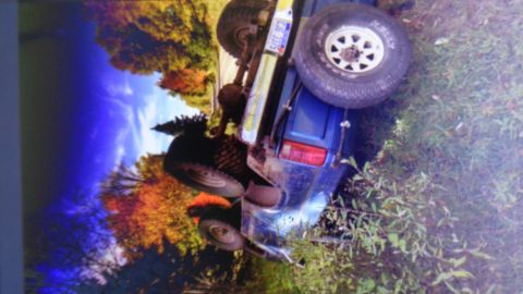 1985 Ford Bronco - A blue SUV is overturned on its side in a grassy area with trees in the background showing fall foliage. The license plate reads "CJB 3375".