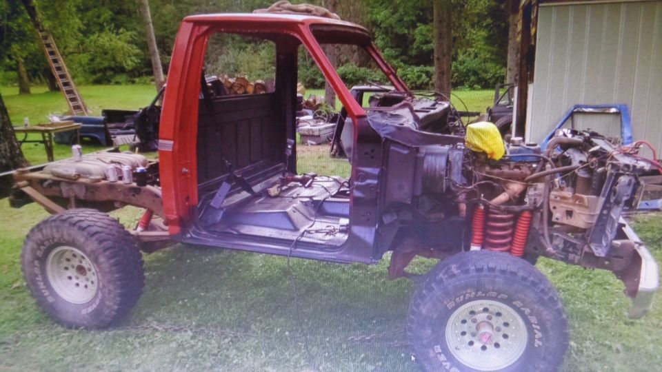Red Ford pickup truck cab and chassis with large off-road tires, disassembled outdoors.