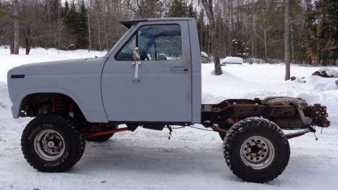 1985 Ford Bronco - Side view of a gray, two-door pickup truck cab with a bare frame and suspension visible. The vehicle is parked in a snowy outdoor setting w…