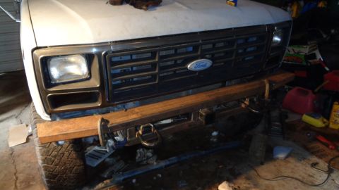 1985 Ford Bronco - The front end of a white Ford Bronco is shown, featuring a custom-built, rustic metal bumper with a tow ring. The vehicle is in a workshop…