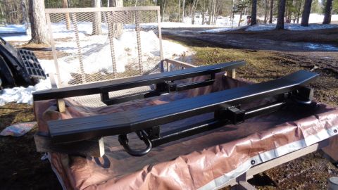 1985 Ford Bronco - Two black painted steel running boards with integrated tow hooks are resting on a tarp-covered surface outdoors. A metal trailer is visible…