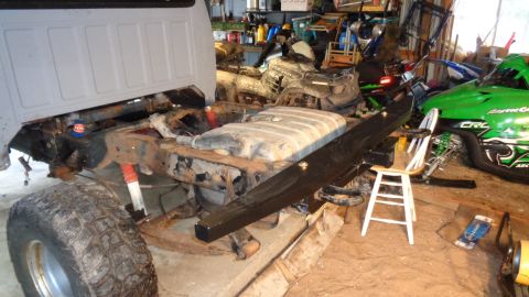 1985 Ford Bronco - The rear frame and suspension of a pickup truck are visible, with a large fuel tank mounted between the frame rails. A black aftermarket bu…