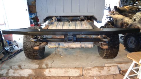 1985 Ford Bronco - The rear of a vehicle chassis is shown, featuring a black aftermarket bumper with a trailer hitch receiver. A large fuel tank is visible be…