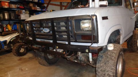 1985 Ford Bronco - The front end of a white Ford Bronco is shown in a garage setting. It has a black aftermarket bumper and grille guard with large off-road t…
