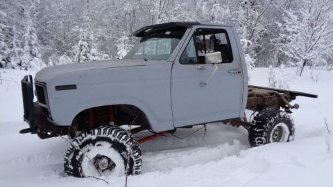1985 Ford Bronco - A light gray, modified pickup truck chassis with large off-road tires is stuck in deep snow in a wooded area. The truck is missing its bed…