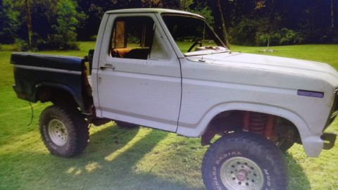 1985 Ford Bronco - A lifted, two-tone pickup truck with a dark green bed and a white cab sits on a grassy lawn. The truck has large off-road tires and appears…