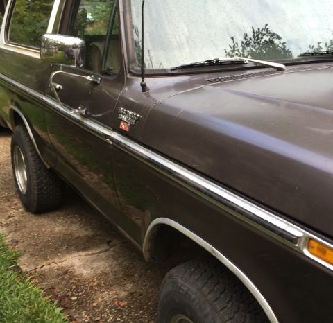 1978 Ford Bronco - Close-up side view of a brown 1978 Ford Bronco with chrome trim and a chrome side mirror. The vehicle is parked on a dirt and concrete surf…