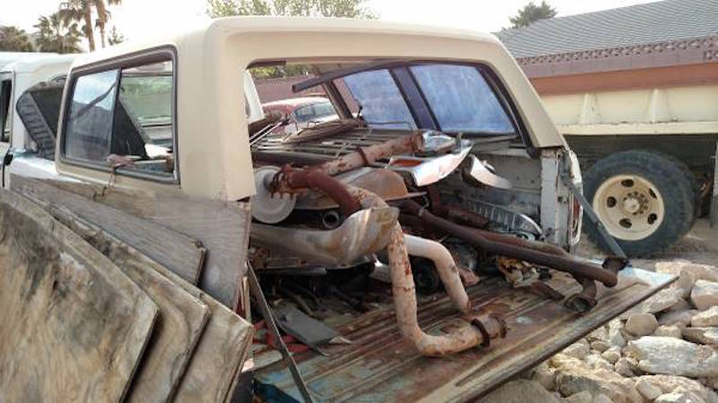 Scrap metal and exhaust parts in the bed of a vintage pickup truck, with a large truck behind it.