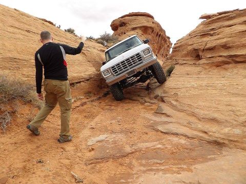 1979 Ford Bronco - A white Ford Bronco is navigating a steep, rocky off-road trail, with a man standing nearby giving a hand signal. The vehicle is tilted at…