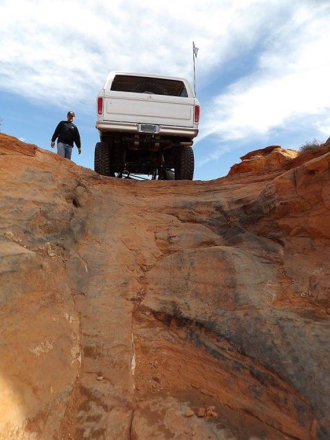1979 Ford Bronco - A white Ford Bronco is shown from the rear, climbing a steep, rocky, off-road trail under a partly cloudy sky. A man in a black jacket and…