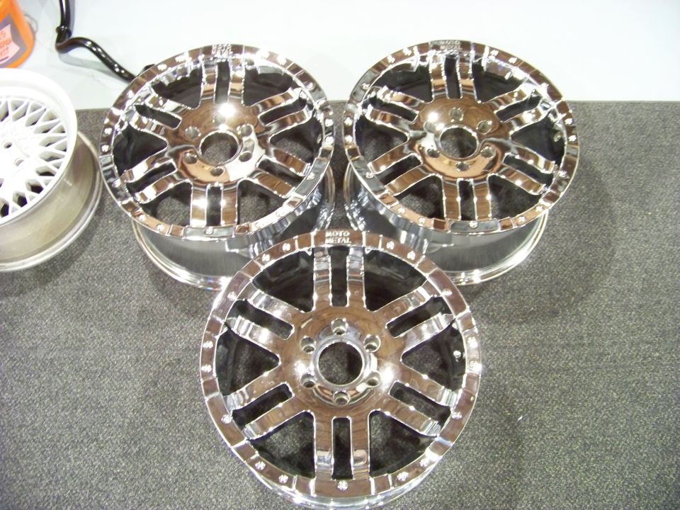 Three shiny chrome aftermarket wheels and one white mesh wheel on a gray carpet.