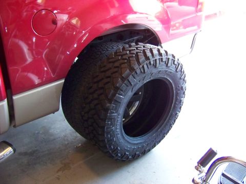 2005 Ford F-150 - A close-up view of a large, aggressive off-road tire mounted on a black rim, positioned next to the rear fender of a red pickup truck. The…