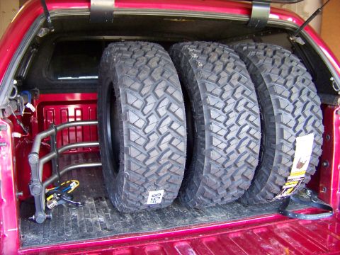 2005 Ford F-150 - Three large off-road tires are stored upright in the bed of a red pickup truck, secured by a metal rack and straps. One tire has a white ta…