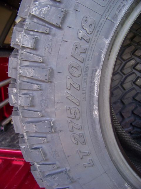 2005 Ford F-150 - A close-up, angled view of a LT 275/70R18 tire mounted on a red vehicle. The tire has aggressive tread blocks and visible text indicating i…