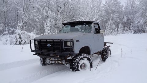 1985 Ford Bronco - A light gray, lifted Ford pickup truck with large, knobby tires is driving through deep snow in a wooded area. The truck has a black brush…