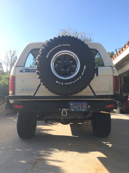 1981 Ford Bronco - The rear view of a beige 1981 Ford Bronco with a large spare tire mounted on the back. The vehicle has a custom rear bumper with a trailer…