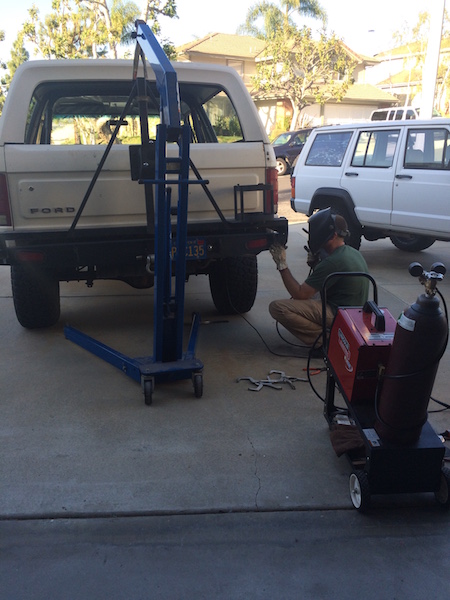 1981 Ford Bronco - A person wearing a welding mask is working on the rear bumper of a light-colored Ford Bronco. A blue engine hoist is positioned behind the…