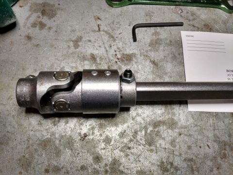 1992 Ford Bronco - A close-up view of a metal universal joint assembly attached to a shaft, resting on a textured metal surface. A small hex wrench and a piec…