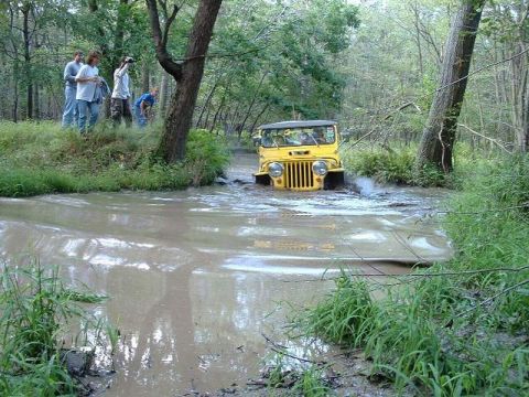 1978 Ford Bronco - A yellow Jeep CJ splashes through a muddy creek bed, with several people watching from the bank.