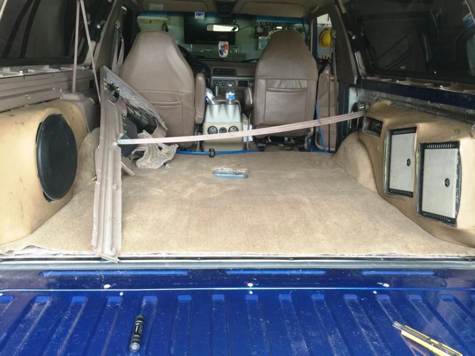 Interior view of a vehicle's cargo area, showing tan carpet, custom speaker boxes on the sides, and front seats