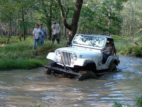 1978 Ford Bronco - A white Jeep CJ navigates through a muddy water obstacle in a wooded area.
