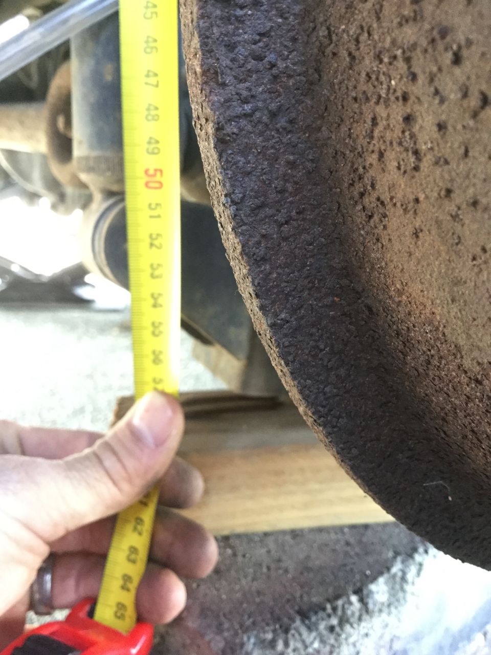 Hand holding a yellow measuring tape against a rusty brake rotor. Tape shows markings from 45 to 65