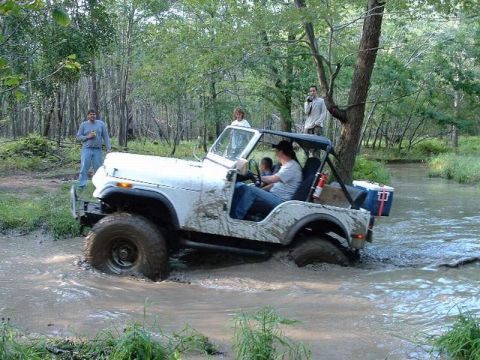 1978 Ford Bronco - A white Jeep CJ-7 navigates a muddy creek in a wooded area, with onlookers present.