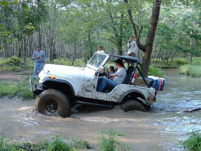 White Jeep CJ-7 with a black soft top driving through muddy water, people watching from the bank