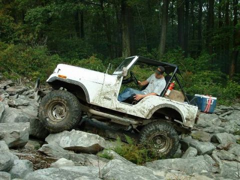 1978 Ford Bronco - A white Jeep CJ-7 tackles a challenging rocky trail with a driver behind the wheel.