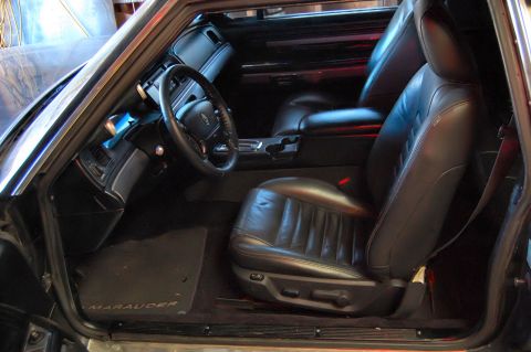 1985 Mercury Grand Marquis - Car interior with black leather seats and dashboard. Floor mat has 'Marauder' text.