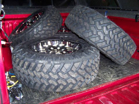 2005 Ford F-150 - Three large off-road tires with chrome rims are loaded into the bed of a red pickup truck.