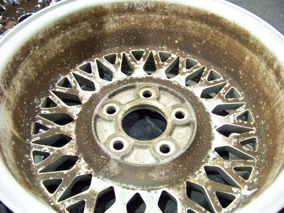 Close-up of a dirty, white alloy wheel with a mesh pattern and five lug nut holes. Text on the wheel reads 'MADE IN ITALY'