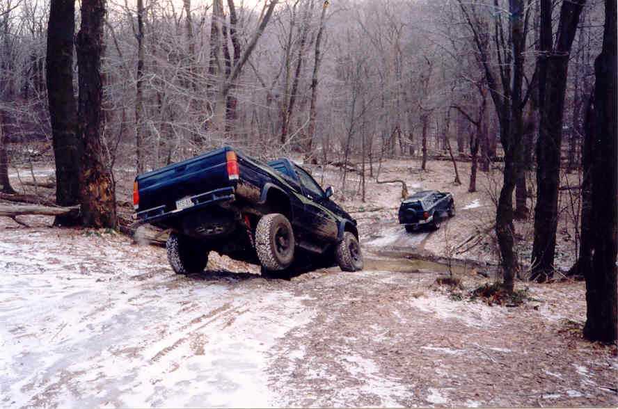 Two dark-colored off-road trucks navigate a muddy and snowy trail through a bare-branched forest