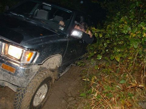 1978 Ford Bronco - A dark-colored SUV navigates a muddy trail at night, with dense foliage encroaching on the vehicle.