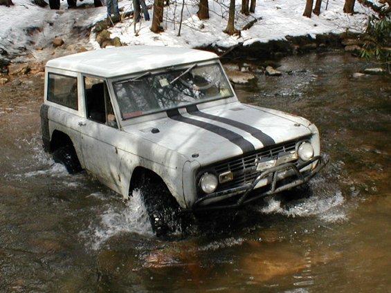White first-generation Ford Bronco with black racing stripes driving through a shallow stream in a wooded area