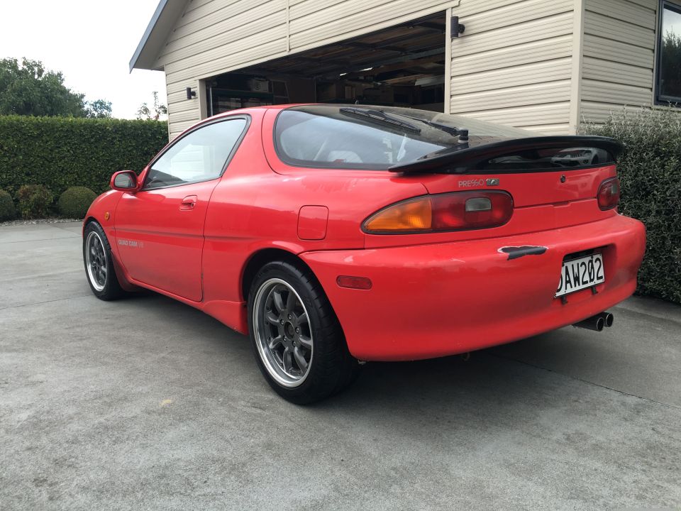 Red 1993 Mazda MX-3 parked in driveway, rear view. Has a spoiler and aftermarket wheels