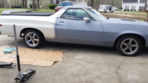 1976 Ford Ranchero - A side view of a Ford Ranchero in primer gray and white, parked on a concrete driveway with a car jack and cardboard on the ground.
