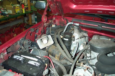 1995 Ford Bronco - Engine bay of a red Ford truck with the hood raised, shot from the driver's front corner in a garage, showing the battery and its red cable…