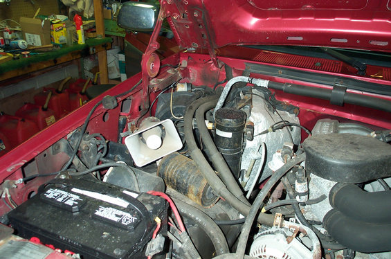 Engine bay of red Ford truck showing battery, distributor with plug wires and alternator