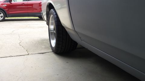 1976 Ford Ranchero - The rear passenger side of a silver vehicle shows two wide tires with chrome rims, parked on a cracked concrete driveway. A red SUV is visi…