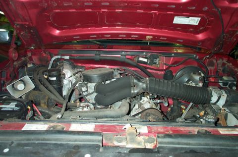 1995 Ford Bronco - Engine bay of a red Ford truck/SUV photographed with the hood up, showing a V8 with black plastic intake tube and air cleaner ducting, brak…