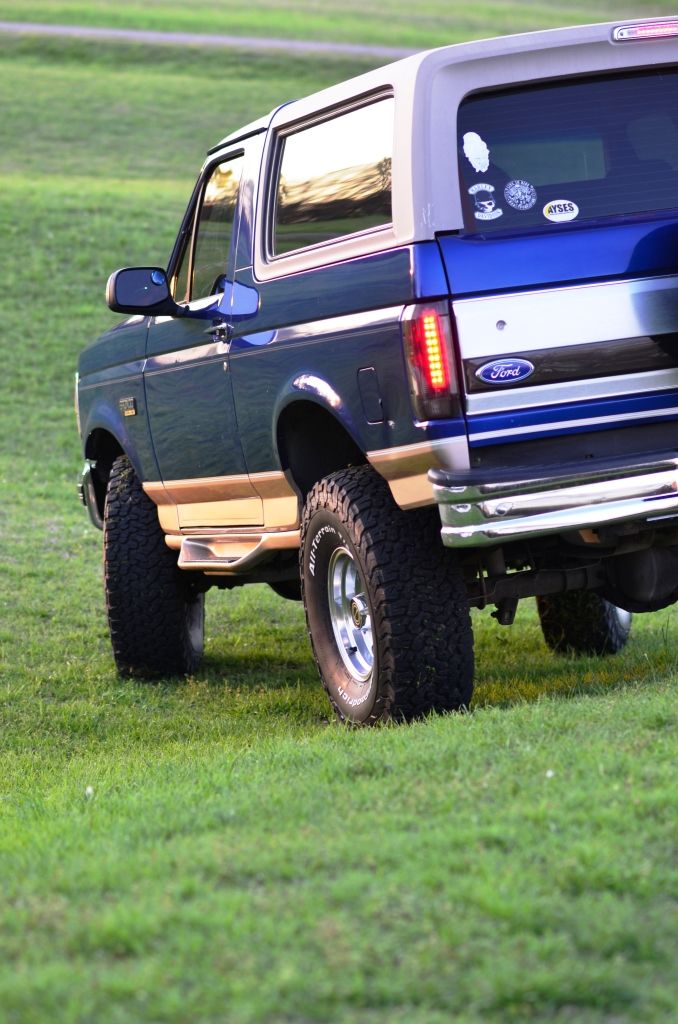 Dark blue and grey Ford Bronco with tan trim, lifted on off-road tires, viewed from rear quarter angle on grassy field